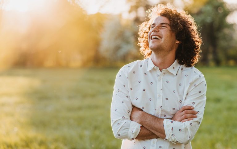BODYMIND CAMPUS glücklicher junger Mann durch bewusstes Stressmanagement Glücklicher junger Mann mit braunen Locken und weißem gemustertem Hemd lächelt in der untergehenden Sonne in den Himmel
