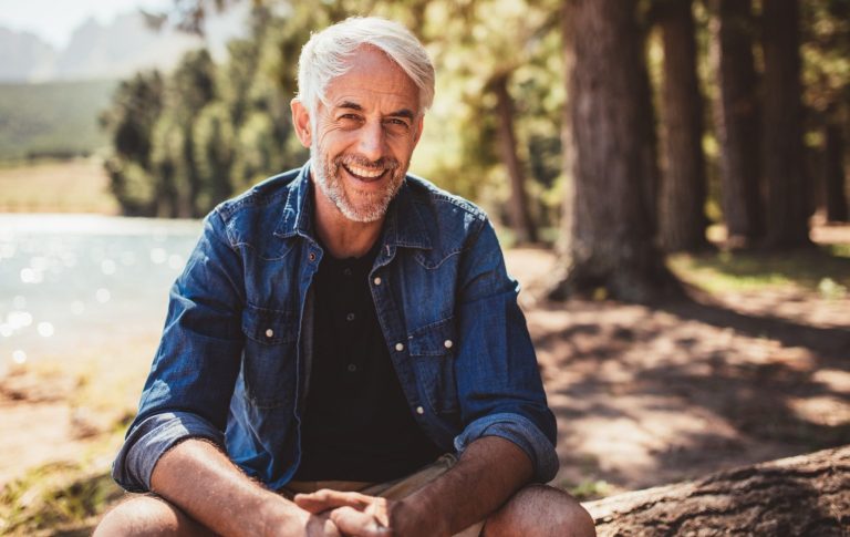BODYMIND CAMPUS glücklicher Mann durch Seminar rund um Achtsamkeit und Resilienz Glücklicher Mann in rustikalem Jeanshemd sitzt in der Natur am See und lächelt in die Kamera