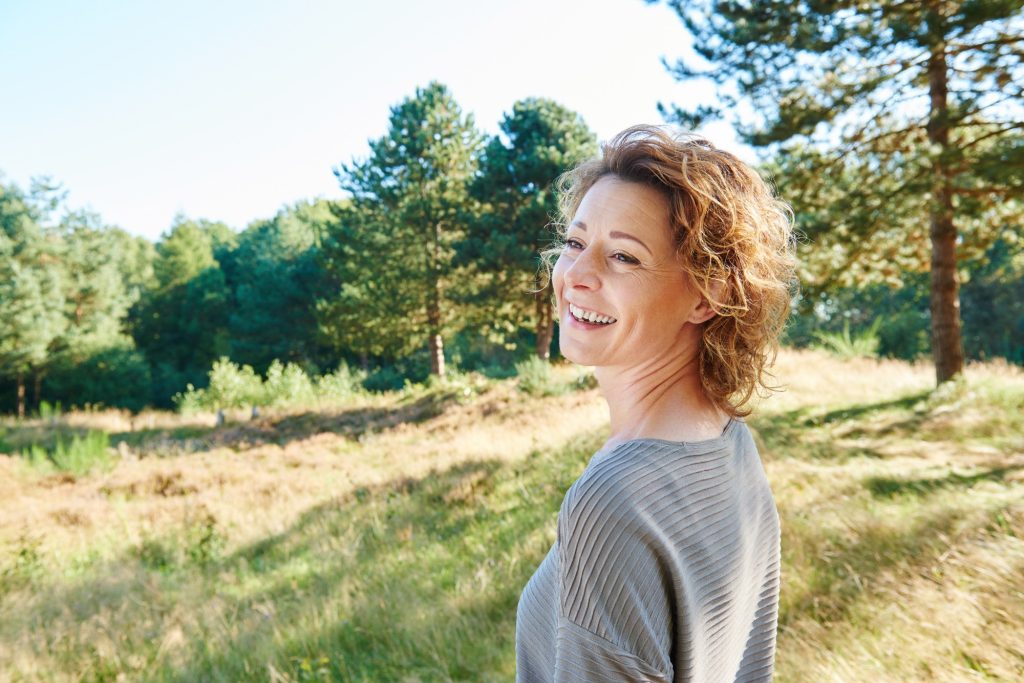 Ältere Frau mit kurzen rötlich-braunen Locken und grauem Pullover, die lächelnd in der sonnigen Natur steht